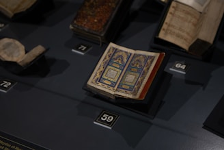 An open, intricately designed book with blue and gold patterns is displayed on a dark surface, surrounded by other historical or religious texts in a display setting.