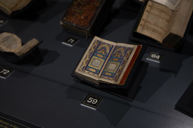 An open, intricately designed book with blue and gold patterns is displayed on a dark surface, surrounded by other historical or religious texts in a display setting.