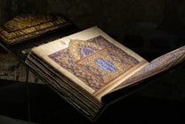 An elaborately decorated manuscript is open, displaying intricate blue and gold geometric patterns and calligraphy on the pages. The cover includes elaborate designs with shades of green and orange.