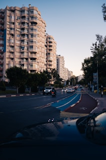 A city street with tall residential buildings on the left side. The sky is clear with a gradient from light blue to darker shades. Vehicles, including a prominent black SUV, are traveling along the road that features a blue bike lane. Trees and greenery line the street, and a few pedestrians are visible on the sidewalk.
