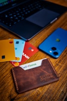 A close-up of a wooden table with a leather cardholder containing several bank cards, each in different colors. A smartphone with a blue case and an Apple logo is positioned next to a laptop with a dark keyboard, partially visible in the background.