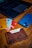 A close-up of a wooden table with a leather cardholder containing several bank cards, each in different colors. A smartphone with a blue case and an Apple logo is positioned next to a laptop with a dark keyboard, partially visible in the background.