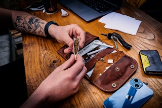 A person with a tattooed arm is rolling a cigarette on a wooden table. There's a leather pouch, some cigarette papers, a set of keys, a smartphone, and some papers scattered around.