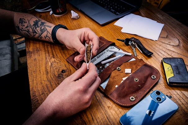 A person with a tattooed arm is rolling a cigarette on a wooden table. There's a leather pouch, some cigarette papers, a set of keys, a smartphone, and some papers scattered around.