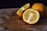 A wooden cutting board holds two whole oranges and a cut orange showing its juicy interior. The surface of the board has visible markings and a dark background enhances the vibrant color of the oranges.