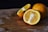 A wooden cutting board holds two whole oranges and a cut orange showing its juicy interior. The surface of the board has visible markings and a dark background enhances the vibrant color of the oranges.