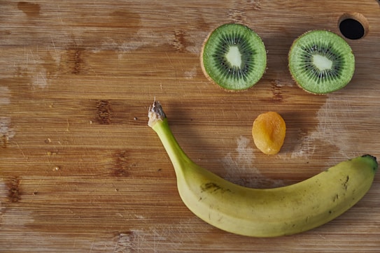 A wooden cutting board features a playful arrangement of fruit. There are two halves of a kiwi fruit positioned as eyes, an orange dried apricot as a nose, and a banana forming a smiling mouth.