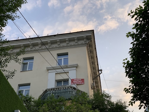 A view of the upper floors of a light-colored building with multiple windows and ornate architectural details. A red and white 'For Sale' sign with contact information is visible on the building's facade. The building is surrounded by greenery, and there are overhead utility lines. The sky above is partly cloudy with patches of blue.
