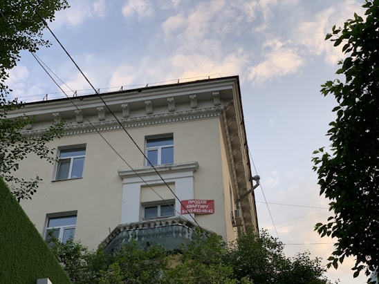 A view of the upper floors of a light-colored building with multiple windows and ornate architectural details. A red and white 'For Sale' sign with contact information is visible on the building's facade. The building is surrounded by greenery, and there are overhead utility lines. The sky above is partly cloudy with patches of blue.