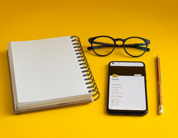 a notepad, glasses, pencil, and cell phone on a yellow surface