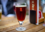 A glass of dark amber beer is placed on a wooden table. The glass has a prominent 'R' logo printed on it. In the background, there is a blurred image of another beer glass and some menu cards.