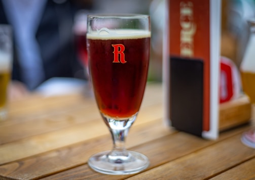 A glass of dark amber beer is placed on a wooden table. The glass has a prominent 'R' logo printed on it. In the background, there is a blurred image of another beer glass and some menu cards.