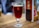 A glass of dark amber beer is placed on a wooden table. The glass has a prominent 'R' logo printed on it. In the background, there is a blurred image of another beer glass and some menu cards.