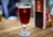 A glass of dark amber beer is placed on a wooden table. The glass has a prominent 'R' logo printed on it. In the background, there is a blurred image of another beer glass and some menu cards.