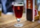 A glass of dark amber beer is placed on a wooden table. The glass has a prominent 'R' logo printed on it. In the background, there is a blurred image of another beer glass and some menu cards.