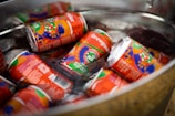 Several colorful, artistically designed drink cans, identifiable as Brewgooder, are submerged in a metal container filled with ice. The cans display vibrant artwork with a mix of orange, green, purple, and blue shades.