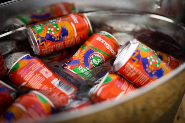 Several colorful, artistically designed drink cans, identifiable as Brewgooder, are submerged in a metal container filled with ice. The cans display vibrant artwork with a mix of orange, green, purple, and blue shades.