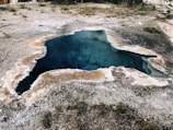 A geothermal hot spring located in a rocky area with mineral deposits surrounding the water. The water appears deep with a vibrant blue color, and the edges of the spring have a crusty white and beige mineral buildup. Sparse vegetation is visible around the spring, and the land is mostly barren and arid.
