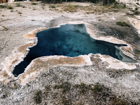 A geothermal hot spring located in a rocky area with mineral deposits surrounding the water. The water appears deep with a vibrant blue color, and the edges of the spring have a crusty white and beige mineral buildup. Sparse vegetation is visible around the spring, and the land is mostly barren and arid.