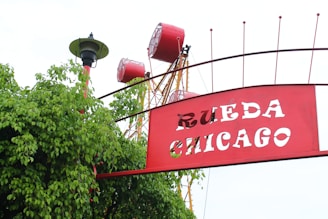 A large red and white sign with the cut-out letters 'RUEDA CHICAGO' is positioned amidst green foliage. Behind the sign, parts of a ferris wheel with red passenger cabins and a yellow hub structure are visible. A classic street lamp with a black base and green canopy is situated to the left.