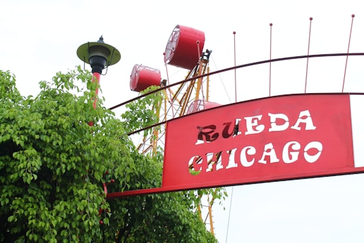 A large red and white sign with the cut-out letters 'RUEDA CHICAGO' is positioned amidst green foliage. Behind the sign, parts of a ferris wheel with red passenger cabins and a yellow hub structure are visible. A classic street lamp with a black base and green canopy is situated to the left.