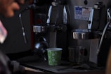 A close-up view of a coffee machine with two coffee cups placed beneath the metallic dispensers. One cup is a colorful paper cup with a leaf pattern, and the other is a transparent glass. The image includes part of a person's blurred face on the left, suggesting interaction with the machine. A shiny, industrial appearance dominates the machine's surface, with labels and buttons visible.