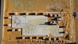 An aerial view of a construction site with a large rectangular foundation surrounded by stacks of materials. The ground is covered in sand, and there are visible tire tracks and footprints across the site.
