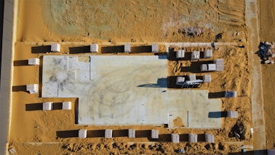 An aerial view of a construction site with a large rectangular foundation surrounded by stacks of materials. The ground is covered in sand, and there are visible tire tracks and footprints across the site.