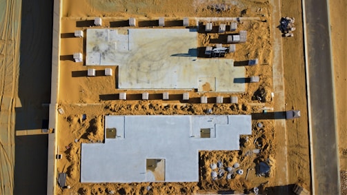 An aerial view of a construction site with two rectangular building foundations on sandy terrain. Numerous stacks of building materials, likely bricks or tiles, are organized around the site. A paved road runs alongside the construction area, and various construction equipment and materials are scattered throughout.