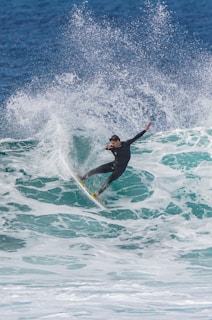 A surfer expertly rides a large wave, with water spraying dramatically around him. The ocean is a vibrant blue and the surfer wears a black wetsuit.