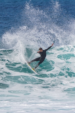 A surfer expertly rides a large wave, with water spraying dramatically around him. The ocean is a vibrant blue and the surfer wears a black wetsuit.