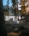 A camping tent is set up in a forested area, surrounded by tall trees with sunlight filtering through. The scene overlooks a serene body of water and distant mountains. Shadows and natural light create a peaceful, secluded atmosphere.