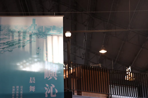 A large poster with a cityscape and harbor in bluish tones is displayed inside a spacious industrial building. The ceiling is high with metal beams and a few hanging lights. The poster includes Chinese characters in prominent font.