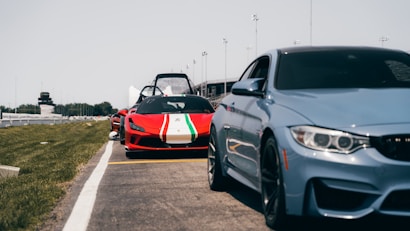 A row of sports cars is lined up on a racetrack with a focus on the red car in the middle bearing a green and white stripe. The sky is clear, and the environment suggests a track event. The foreground features a gray sports car, while fencing, grass, and stadium structures are visible in the background.