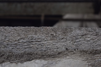 A textured concrete surface with visible roughness and small granules. The background is blurred, suggesting an industrial or urban setting.