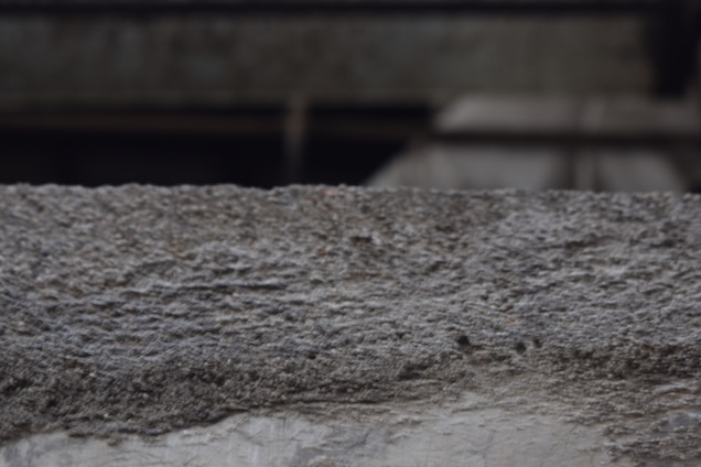 A textured concrete surface with visible roughness and small granules. The background is blurred, suggesting an industrial or urban setting.