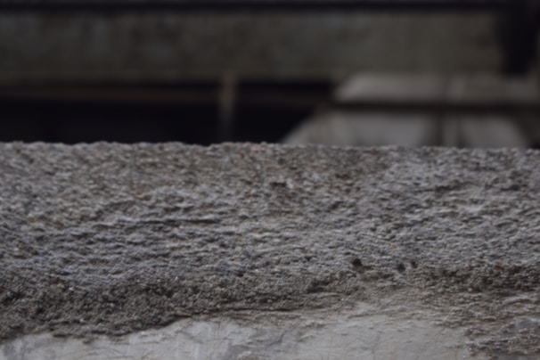 A textured concrete surface with visible roughness and small granules. The background is blurred, suggesting an industrial or urban setting.
