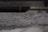 A textured concrete surface with visible roughness and small granules. The background is blurred, suggesting an industrial or urban setting.