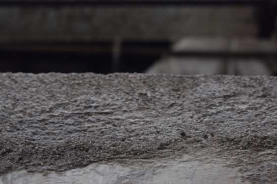 A textured concrete surface with visible roughness and small granules. The background is blurred, suggesting an industrial or urban setting.