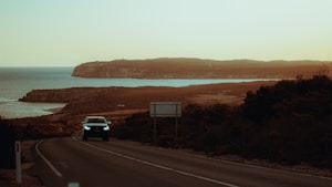 A winding road leads towards a distant coastline, with a car driving along it. The landscape features rugged terrain and cliffs beside a calm sea, under a hazy sky. The setting sun casts a warm glow over the scene.