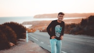 A young man stands laughing on a road with a serene coastal backdrop. The scenery includes a distant shoreline, blue sea, and hilly terrain. The sky is clear, and the scene is bathed in warm sunlight, suggesting a peaceful and cheerful atmosphere.