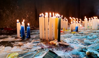 An array of lit candles, predominantly white and blue, stands on a surface covered with melted wax in various colors. The flames flicker against a dark backdrop, creating a warm glow.