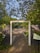 A wooden gate opens to a gravel pathway surrounded by lush greenery and trees with leaves partly shading the ground. A fence with vertical slats lines each side of the gate, and a flowering bush with white blossoms grows nearby. In the distance, another similar gate can be seen further down the path. A sign with a map is mounted on one of the fence panels.