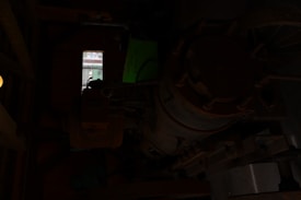 A dimly lit room features an old, rusted mechanical apparatus, possibly part of an industrial machine. A single small window in the background allows limited natural light to enter, and there appears to be a green sign with some text beside the machinery.