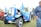 A vintage blue tractor with the brand name Scania displayed on the front is adorned with colorful flags featuring a yellow deer. Other tractors, including a red one with the Volvo logo, are visible in the background. A person in a blue sweater stands to the right, partially cropped out of the frame. The scene is set outdoors on grassy terrain with trees in the distance.
