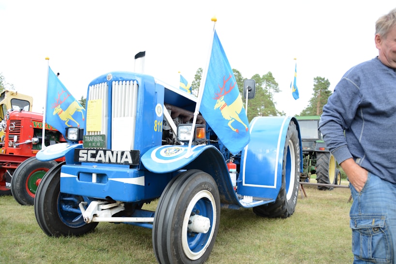 A vintage blue tractor with the brand name Scania displayed on the front is adorned with colorful flags featuring a yellow deer. Other tractors, including a red one with the Volvo logo, are visible in the background. A person in a blue sweater stands to the right, partially cropped out of the frame. The scene is set outdoors on grassy terrain with trees in the distance.