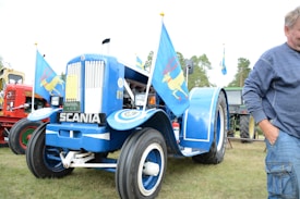 A vintage blue tractor with the brand name Scania displayed on the front is adorned with colorful flags featuring a yellow deer. Other tractors, including a red one with the Volvo logo, are visible in the background. A person in a blue sweater stands to the right, partially cropped out of the frame. The scene is set outdoors on grassy terrain with trees in the distance.