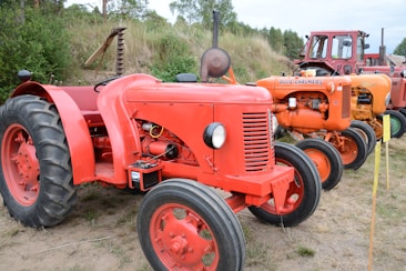 A row of vintage tractors displayed outdoors on a grassy area, with varied designs and colors including red and orange. The tractors are lined up, showcasing their classic and robust build.