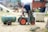 A man is operating a vintage engine attached to a small green and red tractor. Smoke is emanating from the engine. The scene appears to be set outdoors on a sunny day with a crowd of people and more vintage machinery in the background. There is a green cylindrical tank labeled with 'H2O' and 'JR' next to the tractor. Chairs and a canopy are visible, suggesting a leisurely or casual setting.