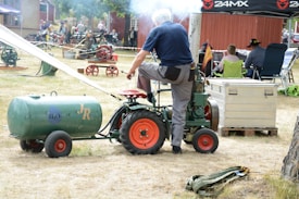 A man is operating a vintage engine attached to a small green and red tractor. Smoke is emanating from the engine. The scene appears to be set outdoors on a sunny day with a crowd of people and more vintage machinery in the background. There is a green cylindrical tank labeled with 'H2O' and 'JR' next to the tractor. Chairs and a canopy are visible, suggesting a leisurely or casual setting.